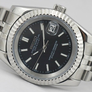 May include: A silver-tone wristwatch with a black dial, a fluted bezel, and a multi-link metal bracelet. The dial features the text SEIKO, OYSTER PERPETUAL DATEJUST, and SUPERLATIVE CHRONOMETER OFFICIALLY CERTIFIED, with a date window showing 21.