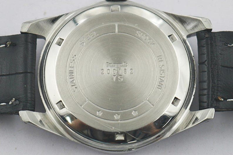 May include: Close-up of the back of a silver-toned wristwatch with a black leather band. The watch face is stainless steel and features the text "STAINLESS STEEL", "SHOCK RESISTANT", and "200182 TS".