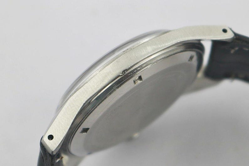 May include: Close-up of a silver-toned wristwatch with a black leather strap. The watch has a clear, domed crystal and a circular face. The watch band is attached to the watch case with small, silver-toned lugs.