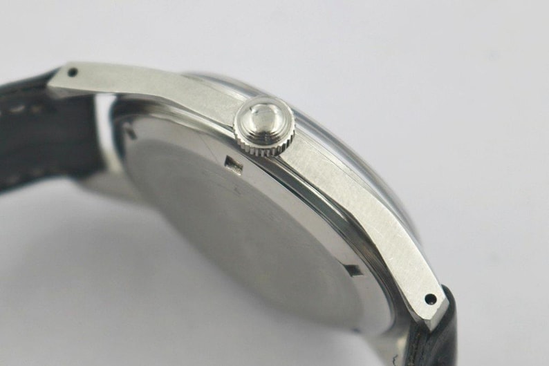 May include: Close-up of a silver wristwatch with a black leather band. The watch has a round face and a textured crown. The watch case is silver and the band is black.