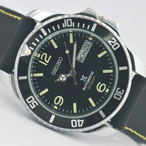 May include: A Seiko automatic wristwatch with a black dial, silver-tone hands, and luminous hour markers. The watch features a rotating black bezel with white numerals, a day-date window at the three o'clock position, and a black silicone strap.