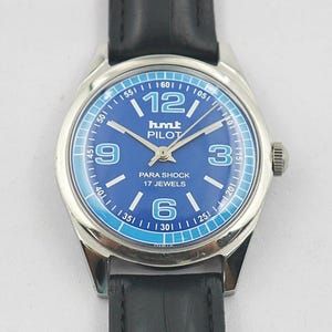 May include: A silver-toned wristwatch with a blue face and black leather band. The watch face features white numerals, hands, and the text "hmt PILOT PARA SHOCK 17 JEWELS". The watch has a round face and a black leather strap.