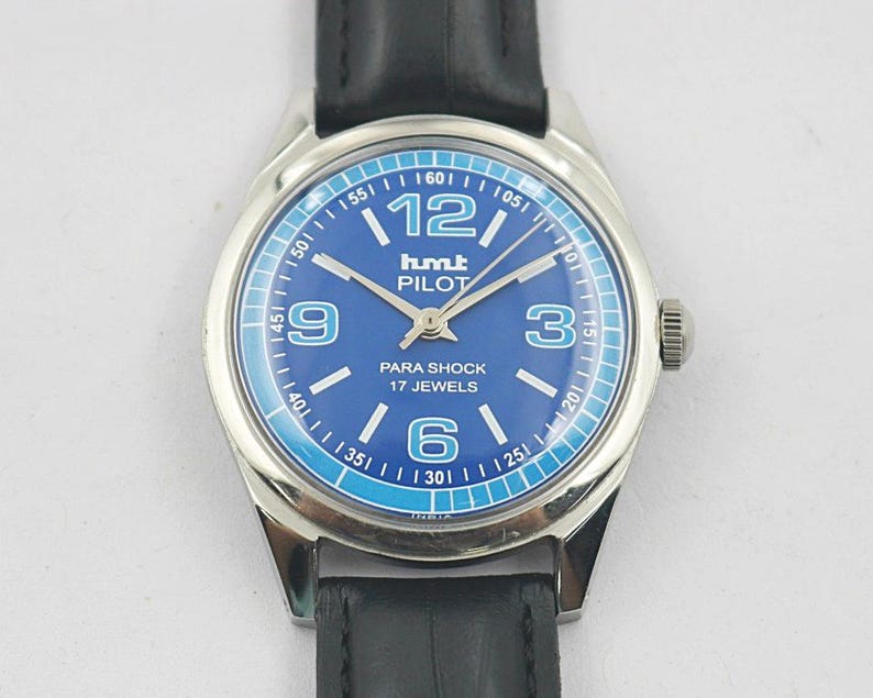 May include: A silver-toned wristwatch with a blue face and black leather band. The watch face features white numerals, hands, and the text "hmt PILOT PARA SHOCK 17 JEWELS". The watch has a round face and a black leather strap.