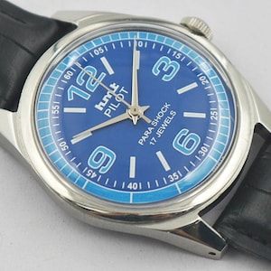 May include: A silver-toned wristwatch with a blue face and black leather band. The watch face features white numerals, the text "hmt PILOT PARA SHOCK 17 JEWELS", and a light blue outer ring. The watch hands are silver.