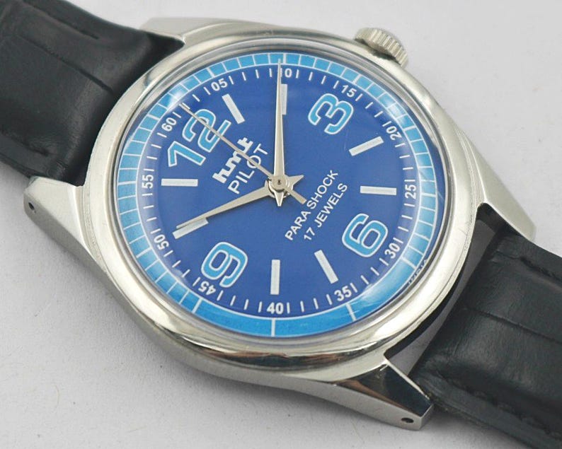 May include: A silver-toned wristwatch with a blue face and black leather band. The watch face features white numerals, the text "hmt PILOT PARA SHOCK 17 JEWELS", and a light blue outer ring. The watch hands are silver.