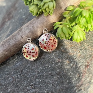 May include: A pair of circular silver pendants featuring a central cross design with red enamel inlay. The surface is decorated with small star and crescent moon motifs arranged symmetrically around the center, each with a small metal loop at the top.