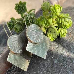 May include: A pair of dangle earrings featuring a circular silver-toned coin charm with a portrait and text, attached to a larger, hammered square-shaped metal plate. The earrings are suspended from long, thin, curved silver-toned wire hooks.