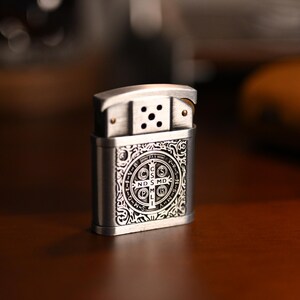 May include: A silver-colored metal lighter with an ornate design. The lighter features a detailed pattern on the front, including a circular emblem with text. The top portion has a hinged lid and a series of holes.