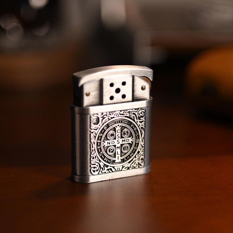 May include: A silver-colored metal lighter with an ornate design. The lighter features a detailed pattern on the front, including a circular emblem with text. The top portion has a hinged lid and a series of holes.