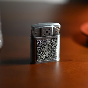May include: A silver-colored metal lighter with an ornate design. The front of the lighter features a detailed emblem with a cross and text. The lighter is rectangular with rounded edges and a hinged top. The background is a blurred wooden surface.