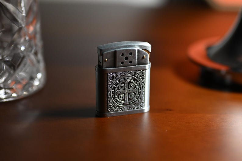 May include: A silver-colored metal lighter with an ornate design. The front of the lighter features a detailed emblem with a cross and text. The lighter is rectangular with rounded edges and a hinged top. The background is a blurred wooden surface.