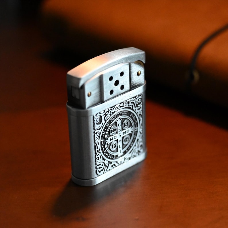 May include: A silver-colored metal lighter with an ornate design. The lighter features a detailed pattern on the front, including a cross and the letters "NDSMD". The top has a hinged lid with a series of holes for the flame.