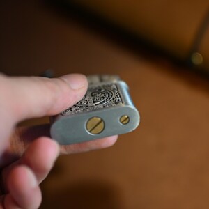May include: Silver-colored metal lighter with ornate engraved detailing on the top. The lighter has a rectangular shape with rounded edges and two brass-colored screws. The lighter is held in a hand.