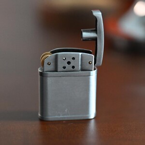 May include: A silver metal lighter with the top open, revealing the internal components. The lighter has a rectangular body and a hinged lid. The flint wheel and other internal parts are visible. The lighter is on a brown surface.