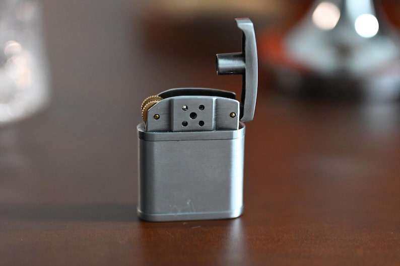 May include: A silver metal lighter with the top open, revealing the internal components. The lighter has a rectangular body and a hinged lid. The flint wheel and other internal parts are visible. The lighter is on a brown surface.