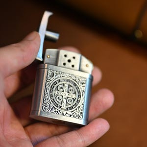 May include: A silver-colored metal lighter with an ornate design. The lighter features a cross-shaped top and a detailed emblem on the front. The emblem includes text and a circular pattern. The lighter is held open in a hand.