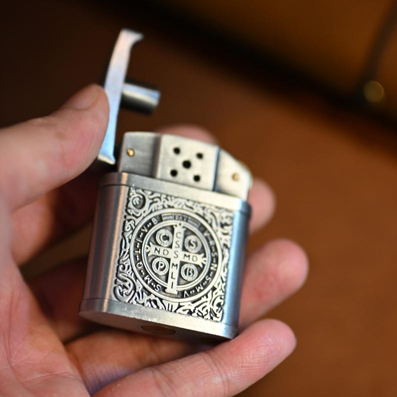 May include: A silver-colored metal lighter with an ornate design. The lighter features a cross-shaped top and a detailed emblem on the front. The emblem includes text and a circular pattern. The lighter is held open in a hand.