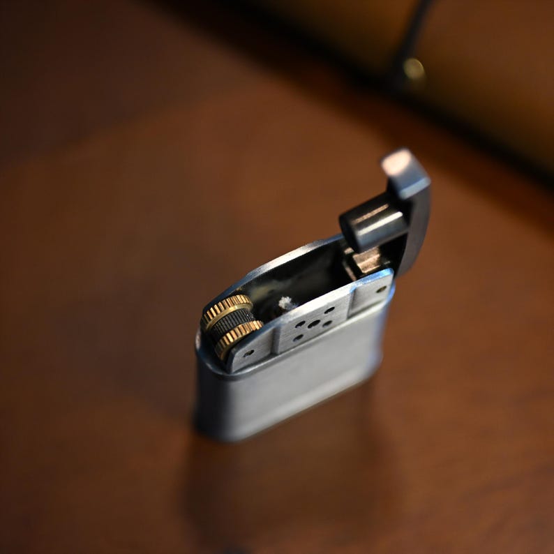 May include: A silver metal lighter with a hinged lid is open, revealing the interior. The lighter has a gold-colored wheel and a black top. The background is a blurred brown surface.