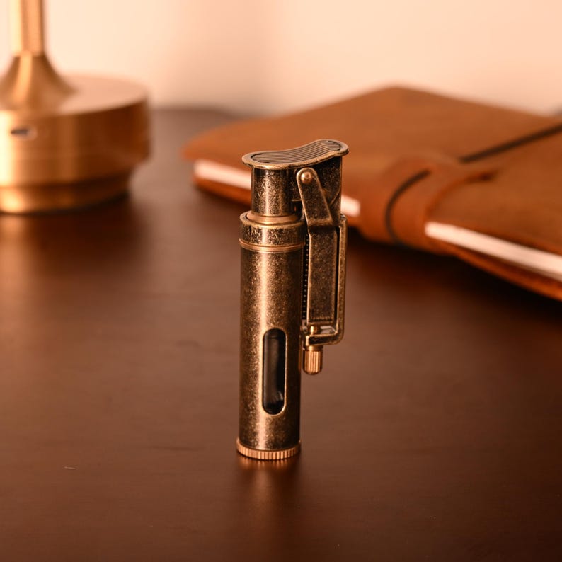 May include: An antique-style lighter with a bronze finish. The lighter has a rectangular body with a hinged top and a side lever. A small window reveals the fuel level. The lighter is on a dark brown surface.