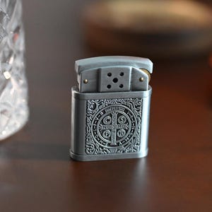 May include: A silver-colored lighter with an ornate, engraved design. The lighter features a rectangular body with a detailed pattern and a hinged top. The design includes a circular emblem with text. The lighter is on a dark brown surface.