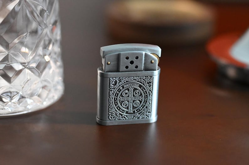 May include: A silver-colored lighter with an ornate, engraved design. The lighter features a rectangular body with a detailed pattern and a hinged top. The design includes a circular emblem with text. The lighter is on a dark brown surface.