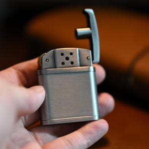 May include: A brushed metal lighter with a hinged top is held open. The lighter has a rectangular body with a textured surface. The top features a series of small holes and is attached to a T-shaped lever. The lighter is held in a hand.