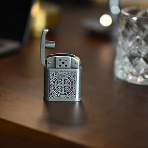 May include: A silver-colored metal lighter with an open top, featuring an ornate design on the front. The lighter is on a dark brown surface, with a clear glass with a diamond pattern in the background.
