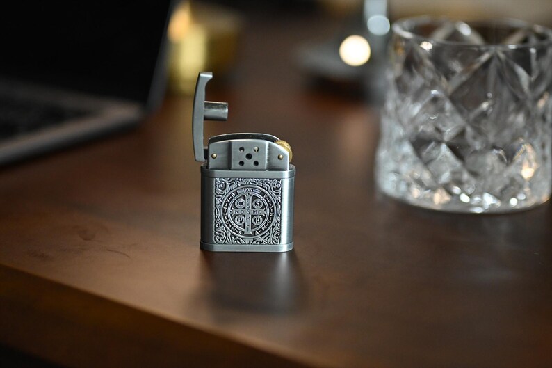 May include: A silver-colored metal lighter with an open top, featuring an ornate design on the front. The lighter is on a dark brown surface, with a clear glass with a diamond pattern in the background.
