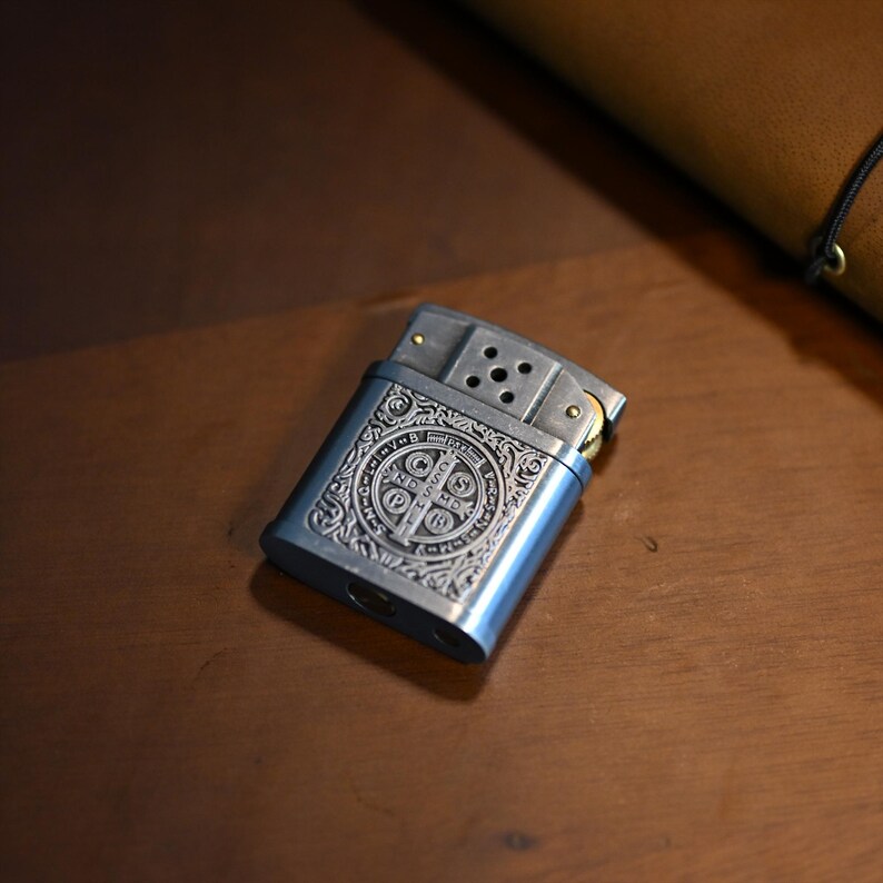 May include: A silver-colored metal lighter with an ornate engraved design. The lighter has a rectangular shape with rounded edges and a hinged top. The design includes a central emblem and decorative scrollwork. The lighter is resting on a wooden surface.