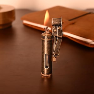 May include: A vintage-style brass lighter with a lit flame. The lighter has a cylindrical body with a viewing window and a hinged mechanism. The background includes a brown notebook and a dark wooden surface.