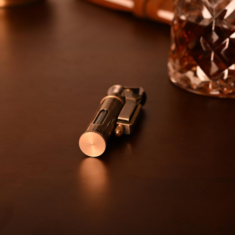 May include: A vintage-style brass lighter with a cylindrical body and a hinged top. The lighter has a small window to view the fuel level. The design is classic and elegant, with a focus on functionality and style. The lighter is on a dark brown surface.