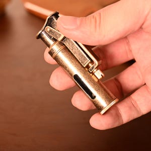 May include: A vintage-style brass lighter with a textured finish. The lighter has a hinged top, a side window, and a small adjustment wheel. The overall design is compact and features a classic, antique aesthetic.