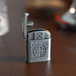 May include: A silver-colored metal lighter with an open lid. The lighter has an ornate design on the front, featuring a circular emblem with text. The background is blurred, with a dark brown surface.