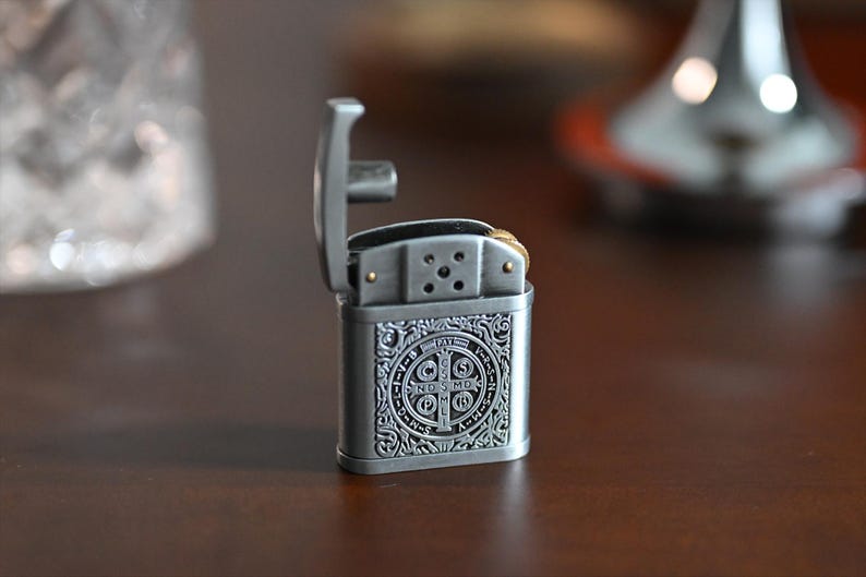 May include: A silver-colored metal lighter with an open lid. The lighter has an ornate design on the front, featuring a circular emblem with text. The background is blurred, with a dark brown surface.