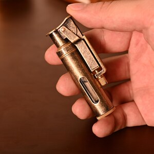 May include: A vintage-style brass lighter with a hinged top and a small viewing window. The lighter has a textured finish and a small, knurled wheel for ignition. The overall design is reminiscent of antique lighters, with a classic, functional aesthetic.