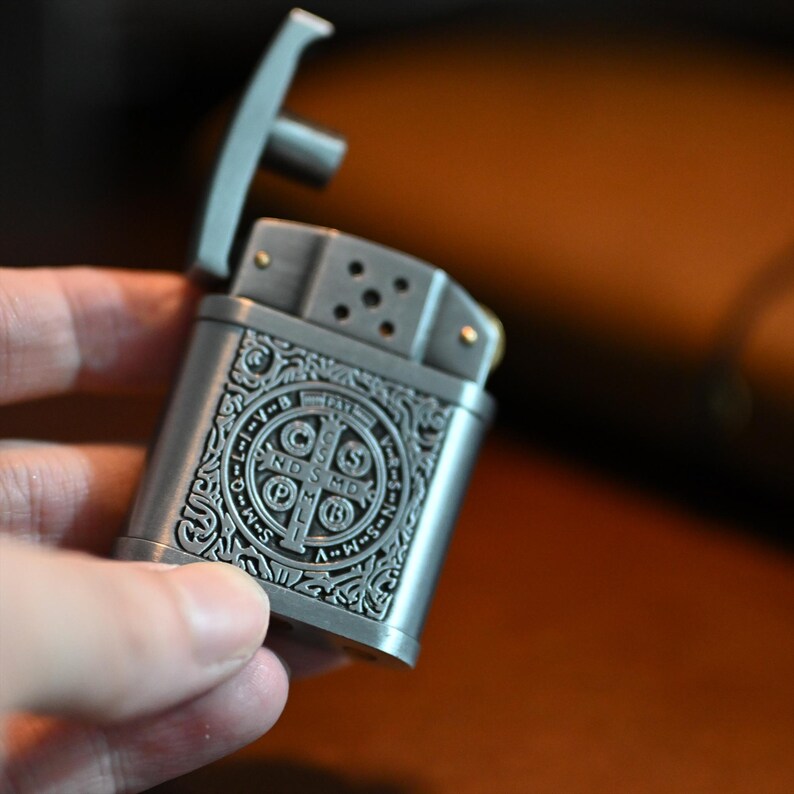 May include: A silver-colored metal lighter with an open lid. The lighter features an ornate design with a cross and the letters "C", "S", "P", "B", "N", "D", "S", and "M". The lighter is held in a hand.