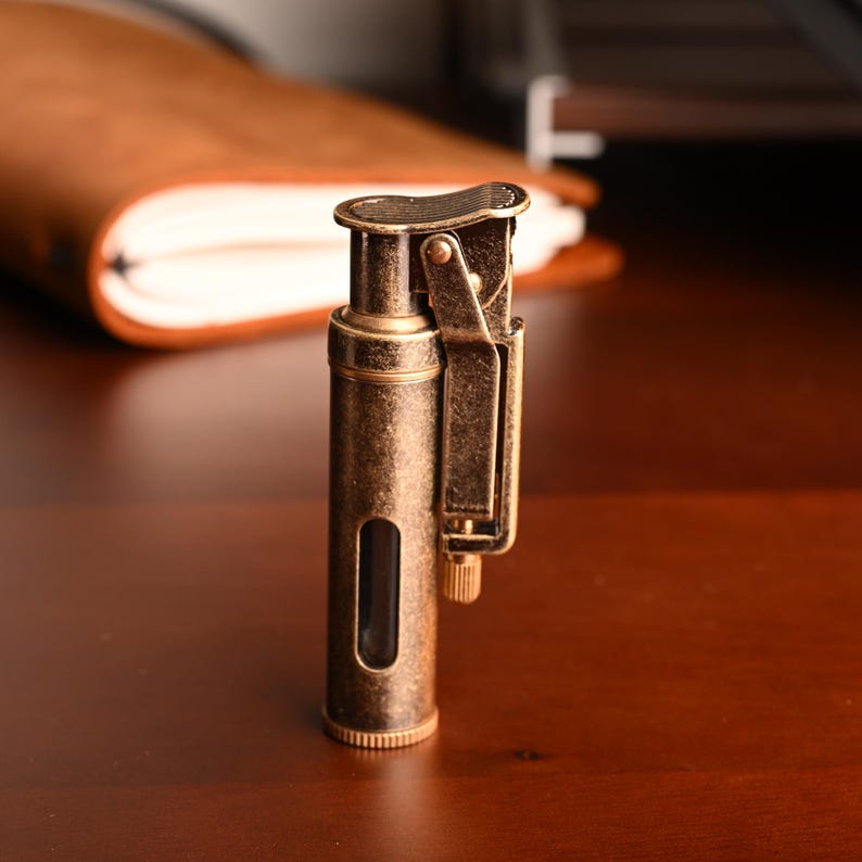 May include: A vintage-style lighter with a bronze finish. The lighter has a rectangular viewing window and a hinged top. The lighter is cylindrical with a textured top and bottom. The lighter is on a dark wooden surface.