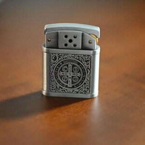 May include: A silver-colored lighter with an ornate design on the front. The design includes a cross and text. The lighter is rectangular with a hinged top and sits on a brown surface.