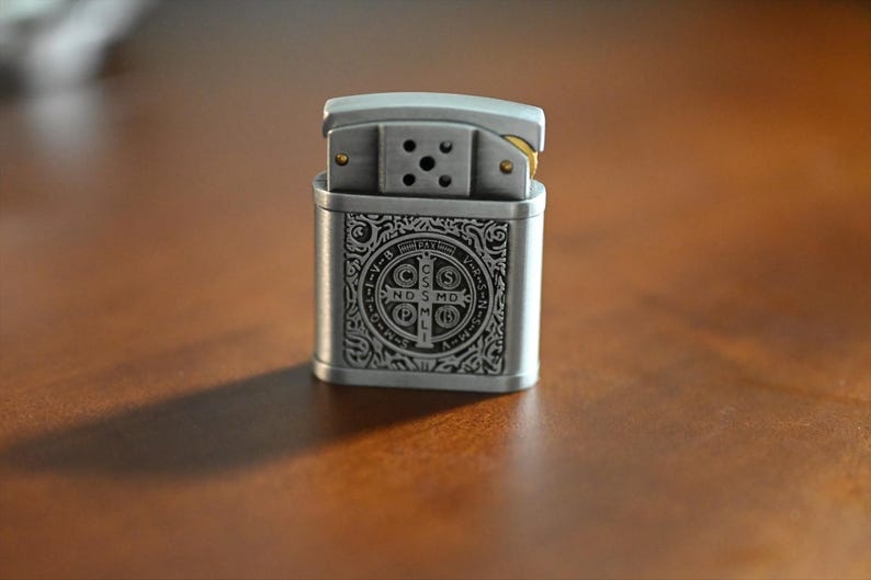 May include: A silver-colored lighter with an ornate design on the front. The design includes a cross and text. The lighter is rectangular with a hinged top and sits on a brown surface.