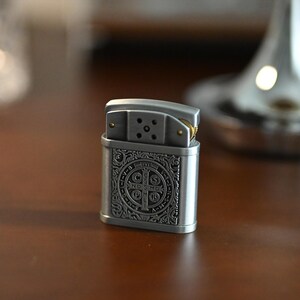 May include: A silver-colored metal lighter with an ornate design on the front. The design features a cross and other symbols. The lighter is sitting on a dark brown wooden surface. The top of the lighter is open, revealing the wick and flint.