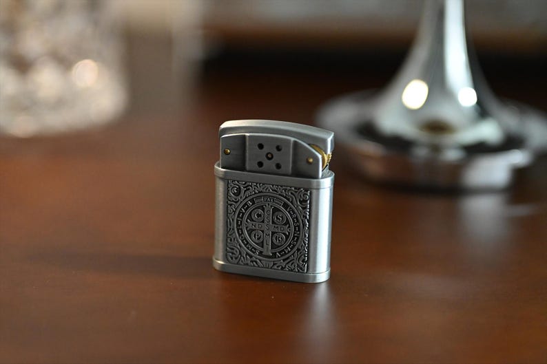 May include: A silver-colored metal lighter with an ornate design on the front. The design features a cross and other symbols. The lighter is sitting on a dark brown wooden surface. The top of the lighter is open, revealing the wick and flint.