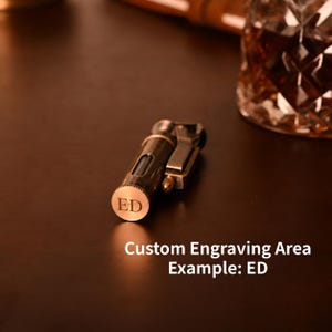 May include: A bronze-colored lighter with the initials "ED" engraved on the bottom. The lighter is on a dark brown surface. The text "Custom Engraving Area Example: ED" is also visible.