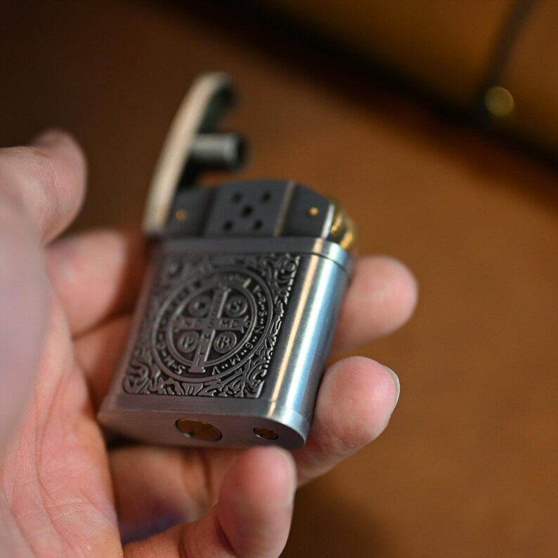 May include: A silver-colored metal lighter with an ornate, engraved design on the front. The design features a cross and circular patterns. The top of the lighter is black, and the hinge is gold-colored. The lighter is held in a hand.