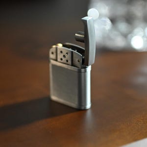 May include: A silver-colored lighter with the top open, revealing the internal components. The lighter is rectangular with rounded edges and sits on a brown surface. A shadow is cast to the left of the lighter.