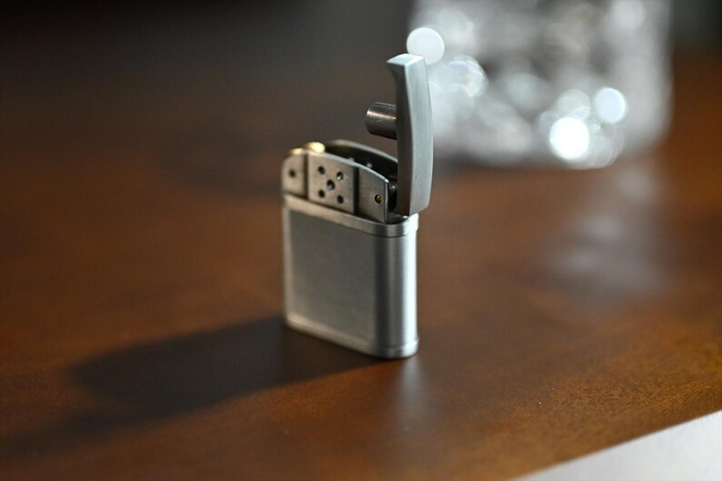 May include: A silver-colored lighter with the top open, revealing the internal components. The lighter is rectangular with rounded edges and sits on a brown surface. A shadow is cast to the left of the lighter.