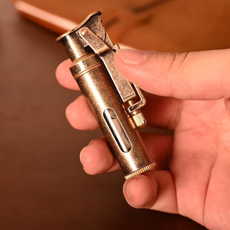 May include: A vintage-style brass lighter with a rectangular fuel window. The lighter has a hinged top and a textured, ridged base. The finish has an aged appearance, suggesting it is an antique or a replica of an older design.