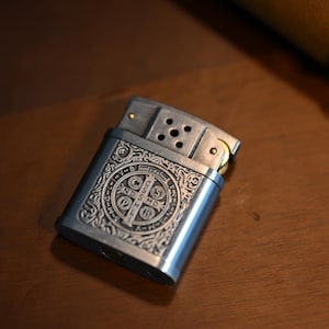 May include: A silver-colored metal lighter with an ornate, engraved design. The design features a central emblem with text and decorative scrollwork. The lighter is rectangular with a hinged top and a visible spark wheel.