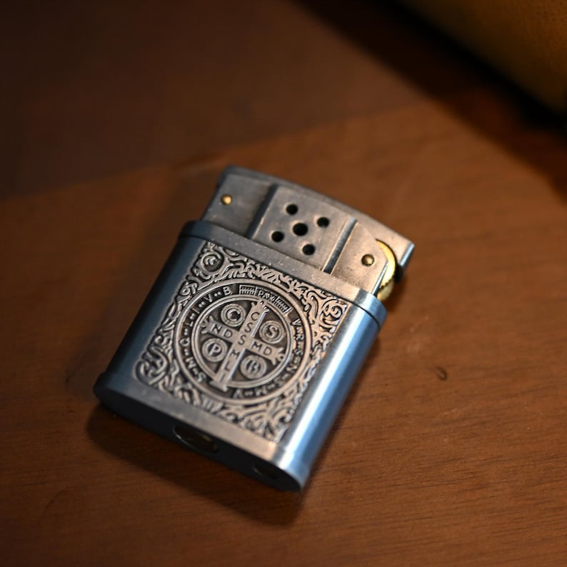 May include: A silver-colored metal lighter with an ornate, engraved design. The design features a central emblem with text and decorative scrollwork. The lighter is rectangular with a hinged top and a visible spark wheel.