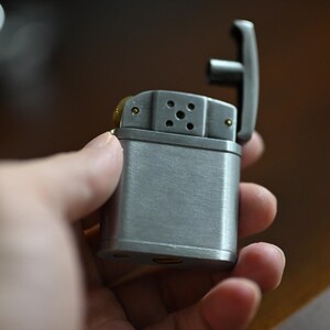May include: A silver-colored lighter with the top open, revealing the internal components. The lighter has a rectangular body and a hinged lid. The lighter is held in a hand.