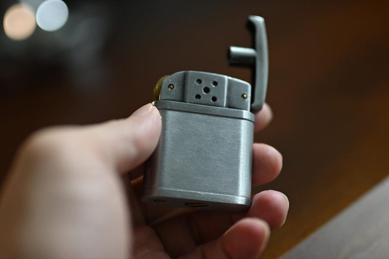 May include: A silver-colored lighter with the top open, revealing the internal components. The lighter has a rectangular body and a hinged lid. The lighter is held in a hand.
